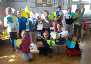 Grupa uczniów - dziewczynki i chłopcy - stoją w sali lekcyjnej i prezentują swoje prace - papierowe serca i gołębie z modlitwami.
